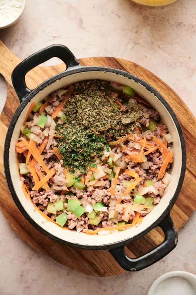 A pot containing uncooked ground meat, chopped vegetables, and herbs sits on a wooden board, ready to be transformed into a flavorful cheeseburger soup.