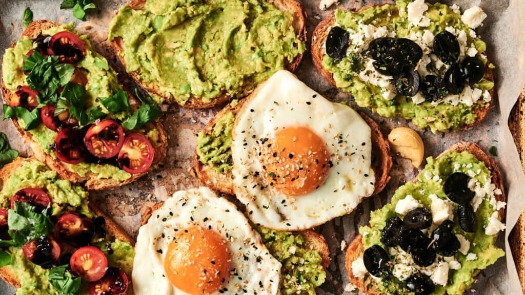 Various avocado toasts topped with cherry tomatoes, fried eggs, black olives, and feta cheese arranged on a parchment-lined surface.