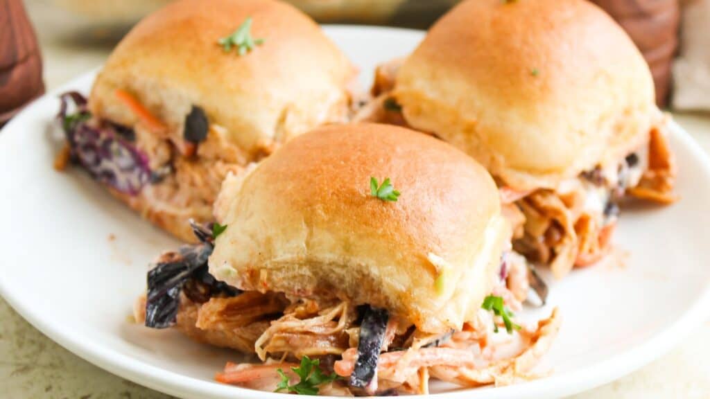 Three pulled pork sliders with coleslaw on a white plate, garnished with a few parsley leaves.