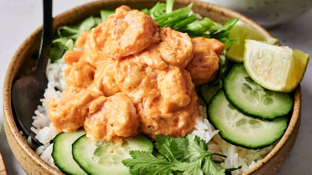 A bowl with creamy shrimp over rice, topped with sliced cucumbers, lime wedges, and fresh cilantro.