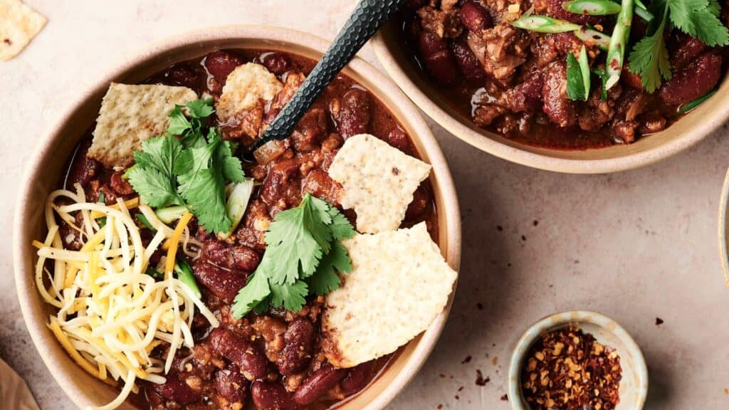 Two bowls of chili with kidney beans and meat, garnished with cilantro, cheese, and tortilla chips. A small bowl of red pepper flakes is nearby.