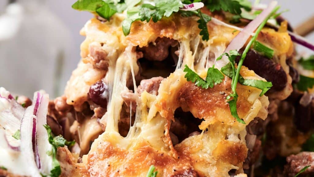 Close-up of a cheesy dish with melted cheese, beef, beans, and scattered cilantro leaves. Red onion slices are visible in the foreground.