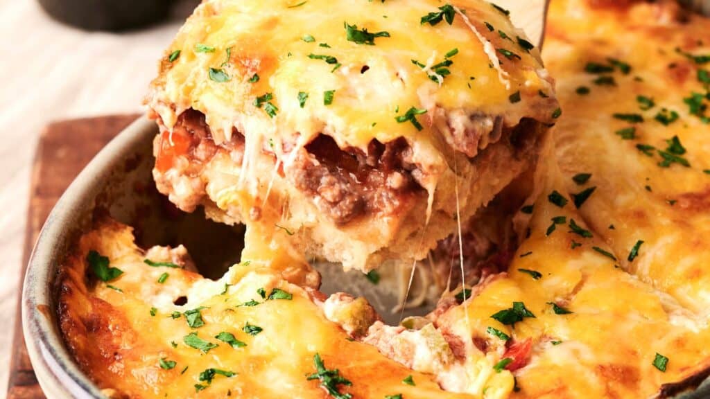 Close-up of a baked lasagna dish with a slice being lifted, revealing layers of meat, sauce, and melted cheese, garnished with herbs.