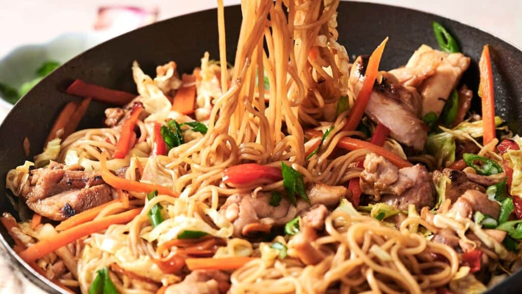 Stir-fried noodles with sliced chicken, carrots, bell peppers, and green onions in a black pan.