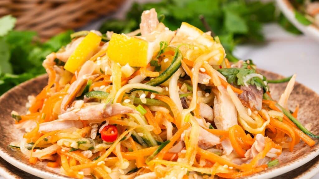 A plate of colorful Asian salad with shredded vegetables, sliced chicken, orange segments, and sesame seeds, garnished with fresh herbs.