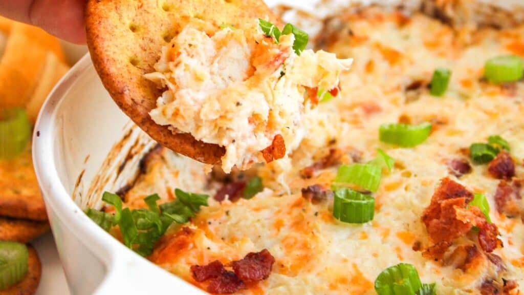 Cracker topped with creamy dip, garnished with chopped green onions and bacon, over a dish filled with the same dip.