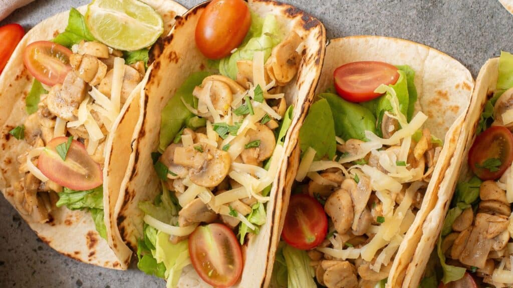 Three soft tacos filled with saut&eacute;ed mushrooms, lettuce, shredded cheese, and cherry tomatoes, garnished with a lime wedge on a gray surface.