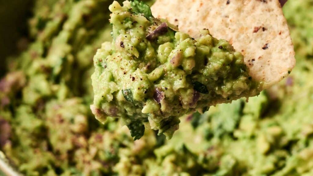 Close-up of a tortilla chip with a scoop of guacamole, showing green avocado, cilantro, and small bits of other ingredients.