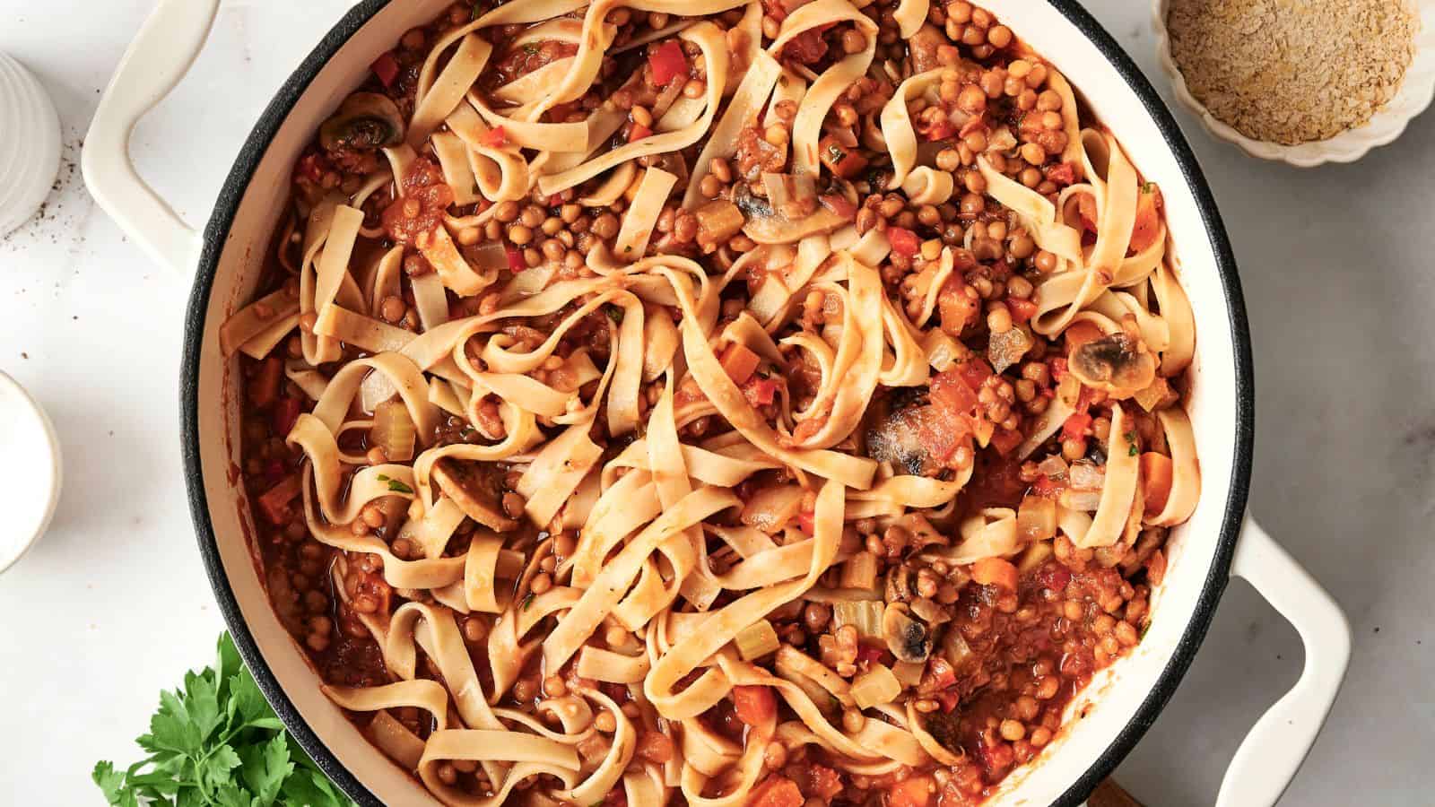A pot of pasta with a tomato-based sauce, lentils, mushrooms, and vegetables on a light surface beside a bowl of grated cheese.