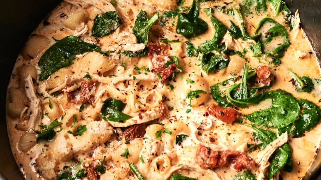 A creamy pasta dish with spinach, shredded chicken, sun-dried tomatoes, shell pasta, and herbs in a sauce, viewed from above.