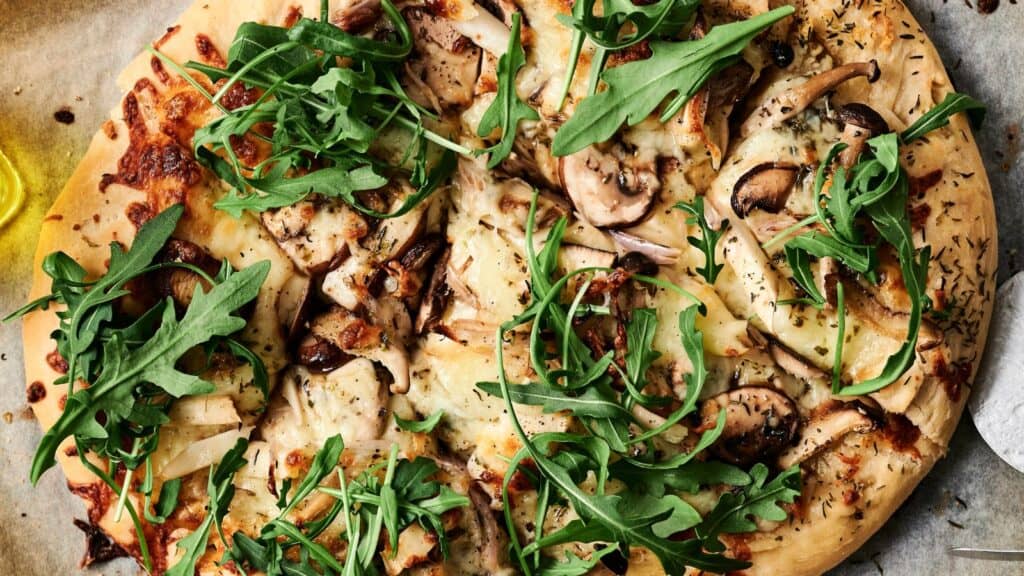 A close-up of a pizza topped with mushrooms, arugula, cheese, and herbs on a wooden surface.