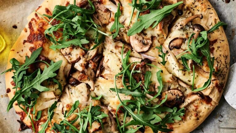 A close-up of a pizza topped with mushrooms, arugula, cheese, and herbs on a wooden surface.