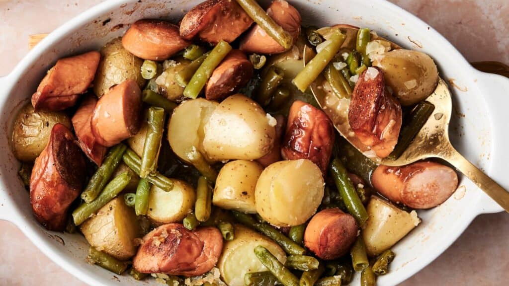 A baked dish with sliced sausages, potatoes, and green beans in a white casserole dish. A golden spoon is partially visible on the right side.