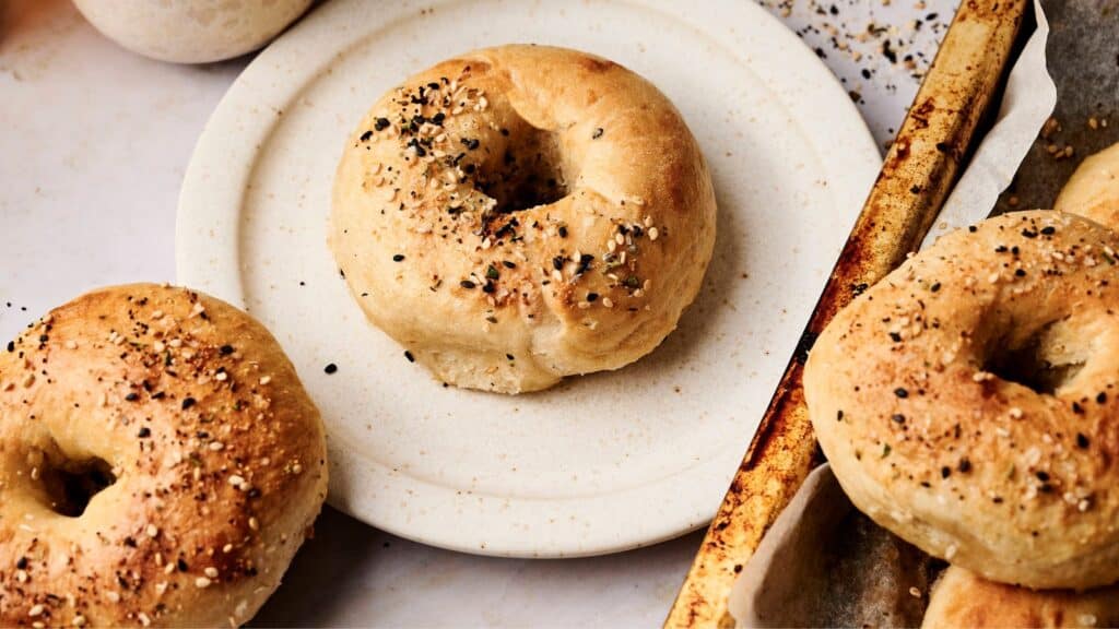 Plate of sesame seed bagels with more bagels on a baking tray nearby.