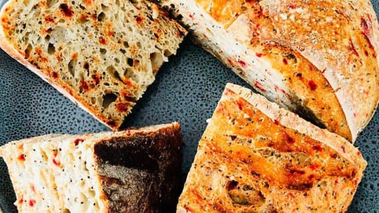 Slices of artisan bread with visible crust and open crumb, featuring specks of red and black seasoning, on a textured dark surface.
