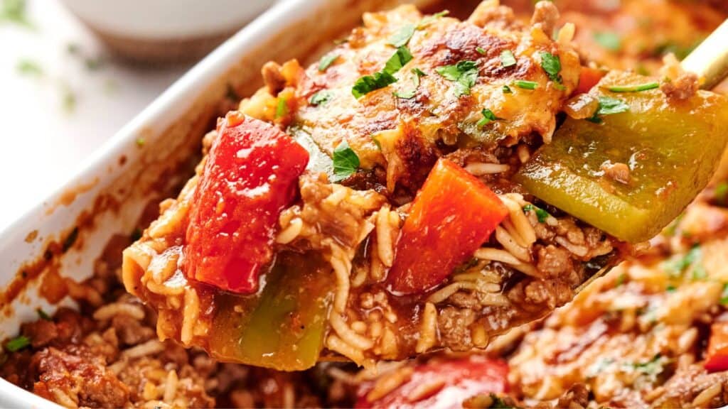 A close-up of a stuffed pepper casserole with ground meat, melted cheese, rice, and red and green bell peppers, garnished with herbs.