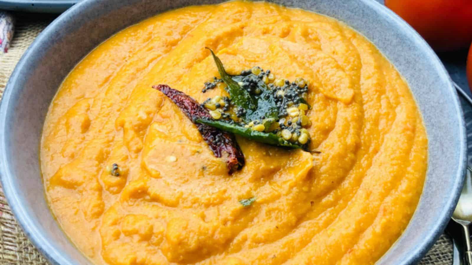 A bowl filled with orange-colored chutney, garnished with mustard seeds, dried chili, and curry leaves.