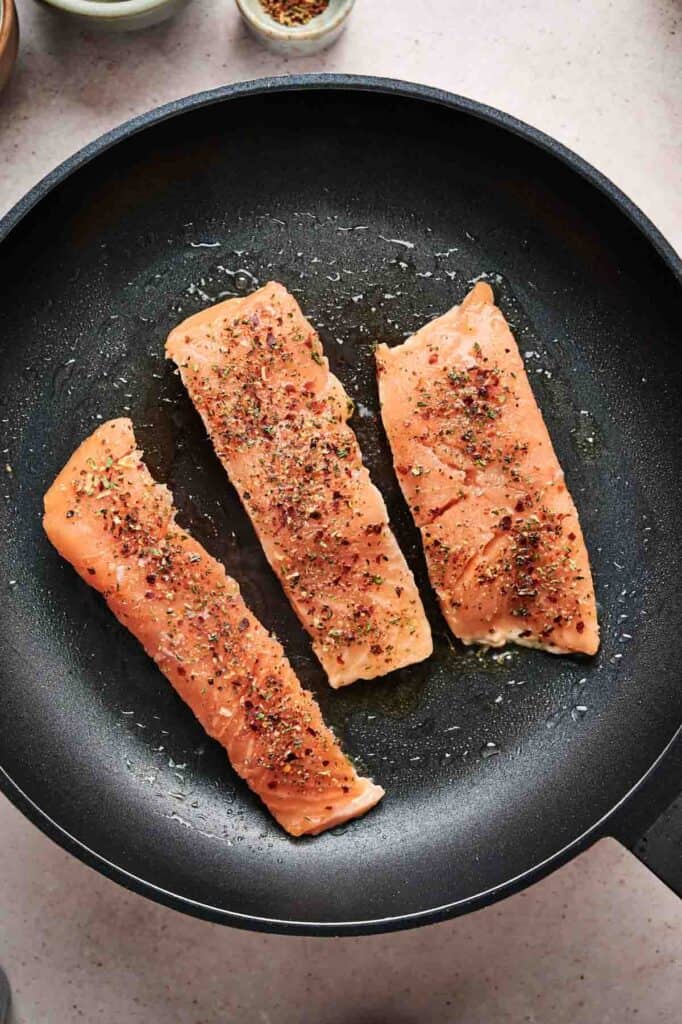 Three seasoned salmon fillets are cooking in a black frying pan with oil.
