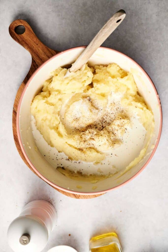 A creamy bowl of mashed potatoes, seasoned with milk and pepper, sits invitingly on a wooden board. A wooden spoon rests inside, ready to serve up comfort in every bite.
