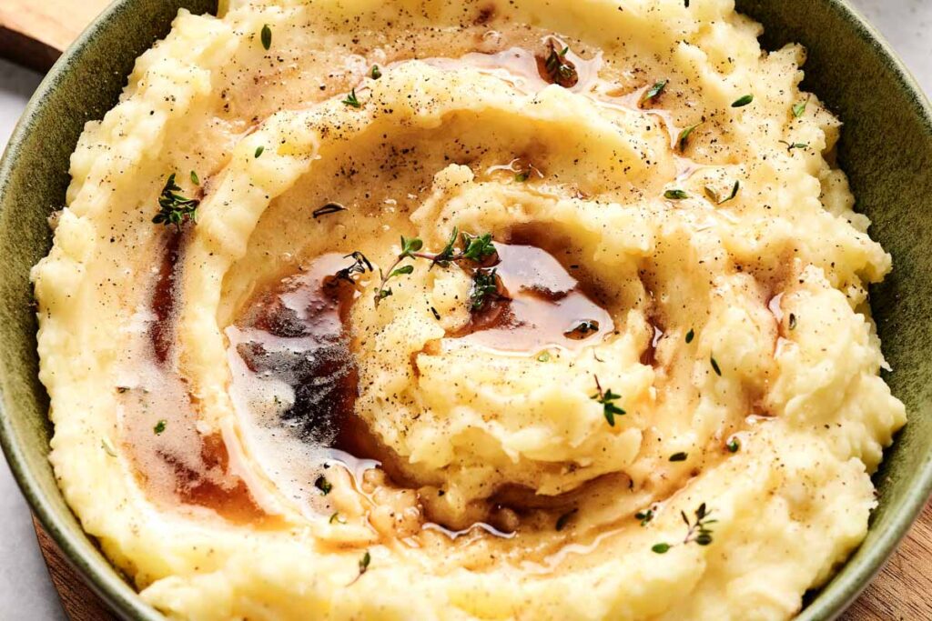 A hearty bowl of mashed potatoes topped with melted butter, black pepper, and fresh herbs.