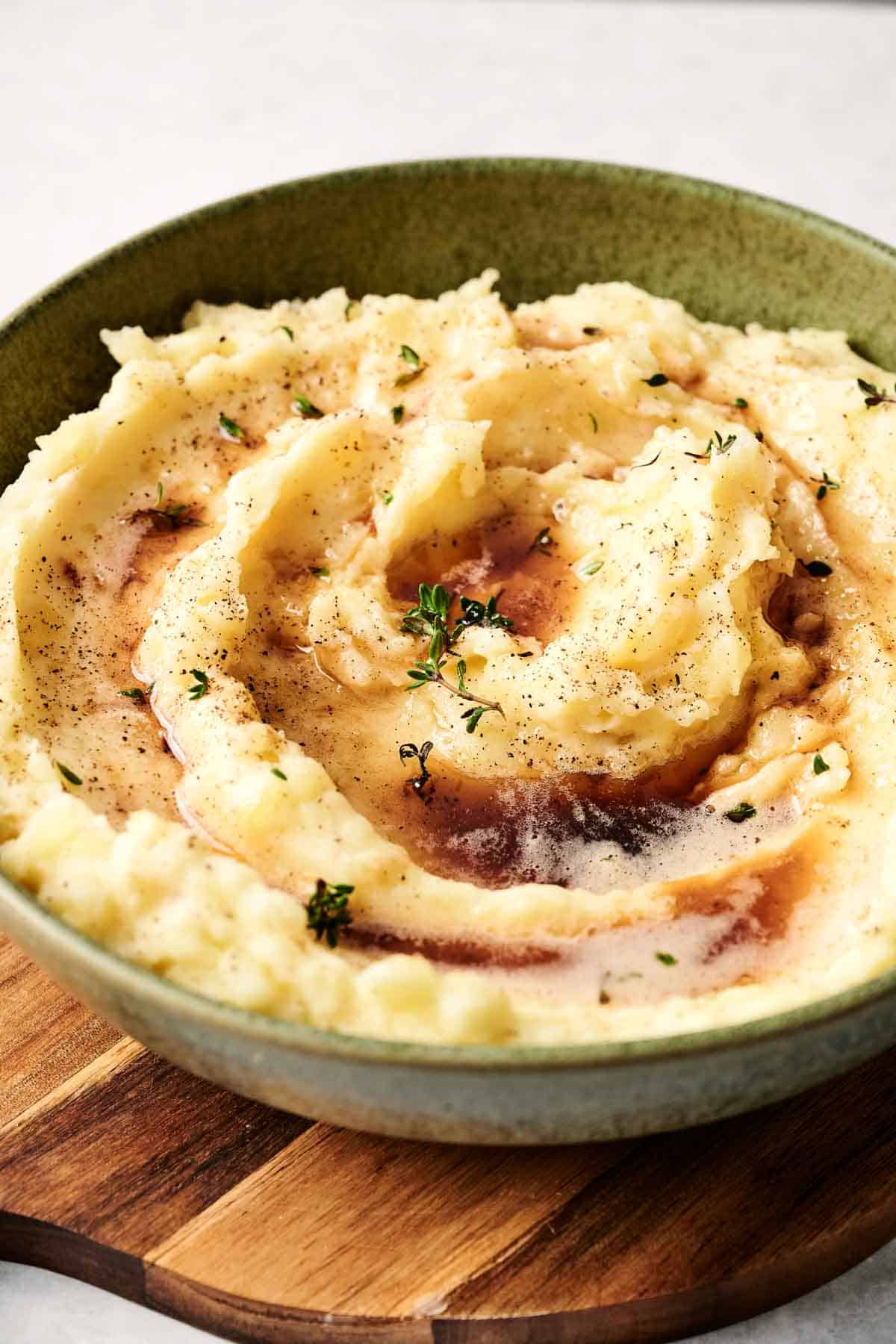 A delightful serving of mashed potatoes, topped with rich gravy and a sprinkle of herbs, elegantly presented in a green bowl on a rustic wooden board.