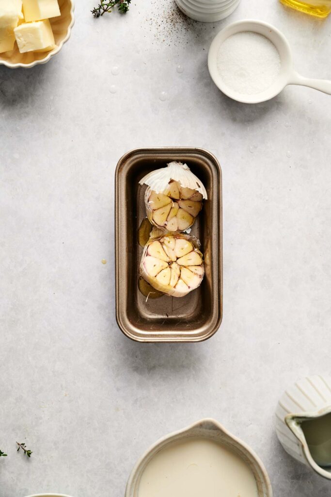 Two halved garlic bulbs with olive oil in a small metal loaf pan, surrounded by butter, cream, sugar, and other ingredients on a white countertop—everything you need to elevate your mashed potatoes to the next level.