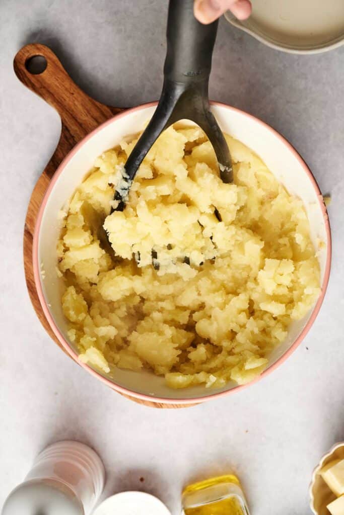 A bowl of creamy mashed potatoes is being prepared with a black masher on a wooden board, surrounded by a salt shaker and oil.