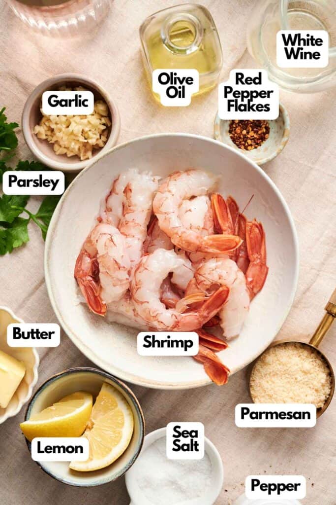 A bowl of raw shrimp, perfect for shrimp scampi, is surrounded by garlic, parsley, butter, lemon, sea salt, olive oil, white wine, red pepper flakes, Parmesan, and pepper on a light cloth background.