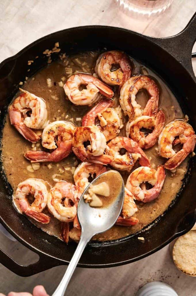 Shrimp scampi sizzles in a skillet as it cooks in a rich brown sauce, with garlic pieces adding a savory touch. A hand skillfully stirs the sauce with a spoon, ensuring each shrimp is perfectly coated and infused with flavor.