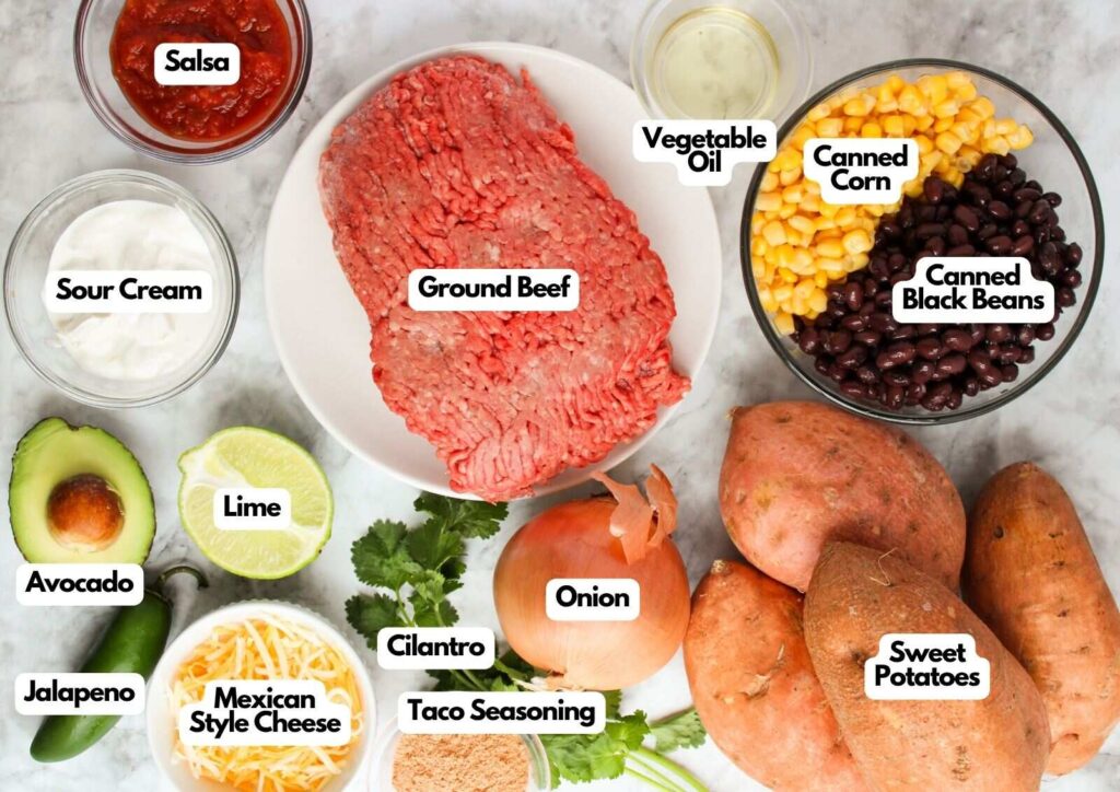 A variety of labeled Southwest Stuffed Sweet Potatoes ingredients are arranged on a counter, featuring ground beef, black beans, corn, avocado, salsa, lime, jalapeño, and more.
