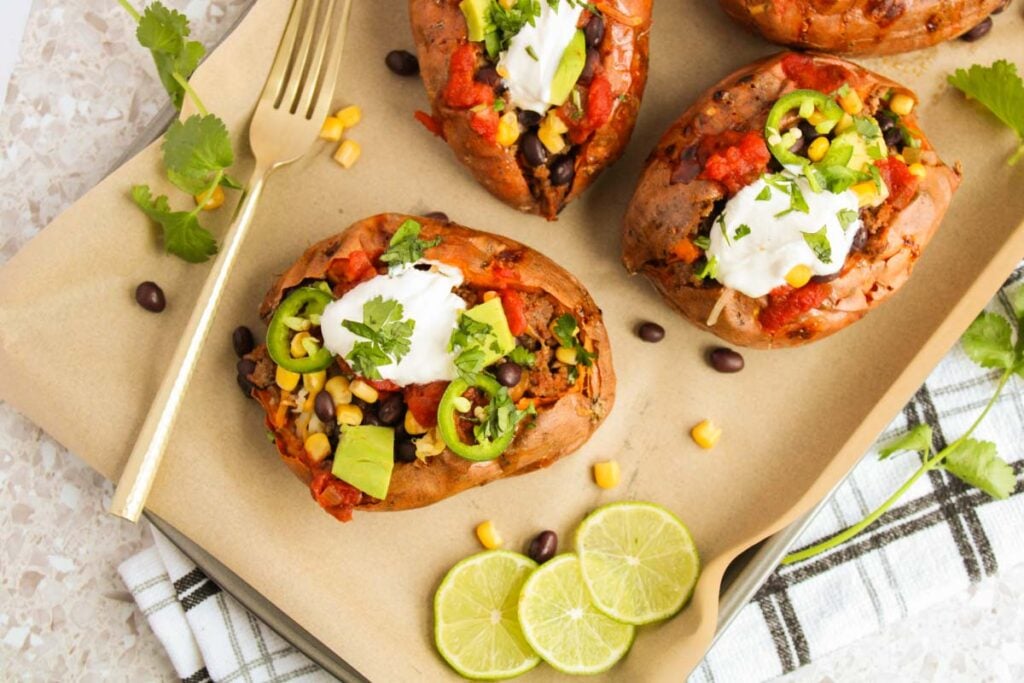 Southwest Stuffed Sweet Potatoes are filled with black beans, corn, diced tomatoes, jalapeño slices, creamy avocado, and topped with sour cream and cilantro. Lime slices and a fork are set alongside for extra flavor.