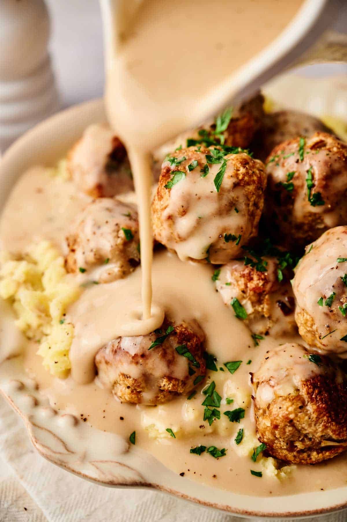 Gravy is being poured over a plate of turkey meatballs and mashed potatoes, garnished with chopped parsley.