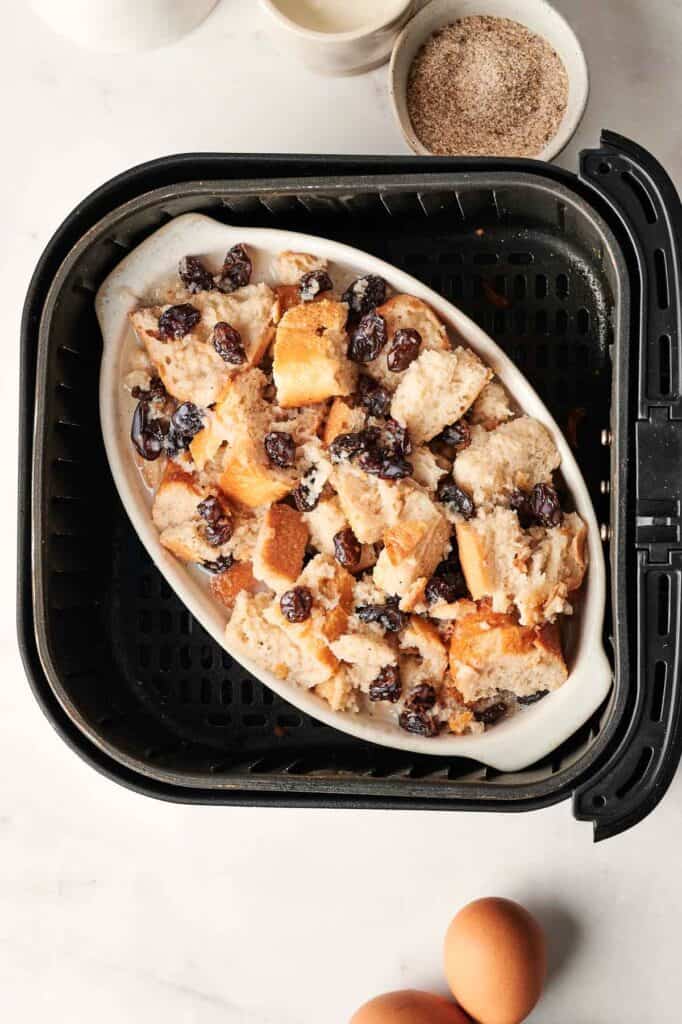 Air Fryer Bread Pudding with raisins sits in the basket, accompanied by two eggs and a bowl of cinnamon sugar, all next to a cup of milk.