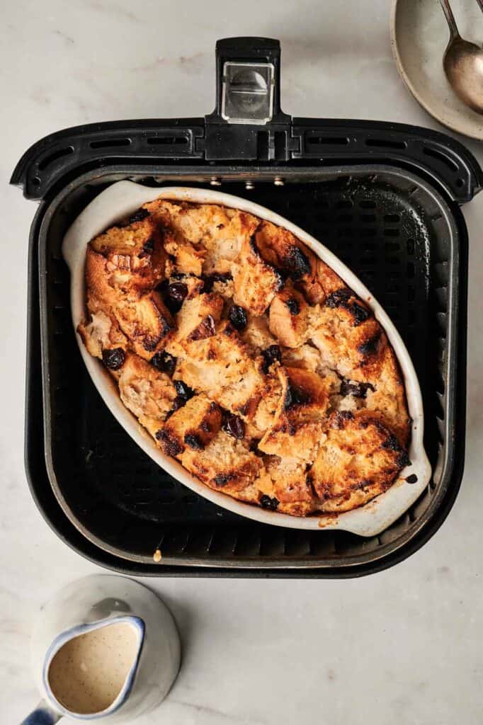 An indulgent air fryer bread pudding awaits in a white oval dish, nestled inside the air fryer. Accompanied by a pitcher of sauce and a spoon, this dessert rests invitingly on the cool marble surface.