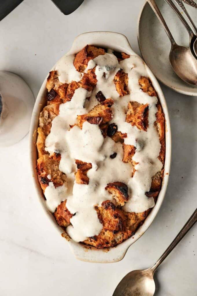 An oval dish of air fryer bread pudding, topped with a luscious white sauce, sits invitingly on the table next to two spoons and an empty plate.