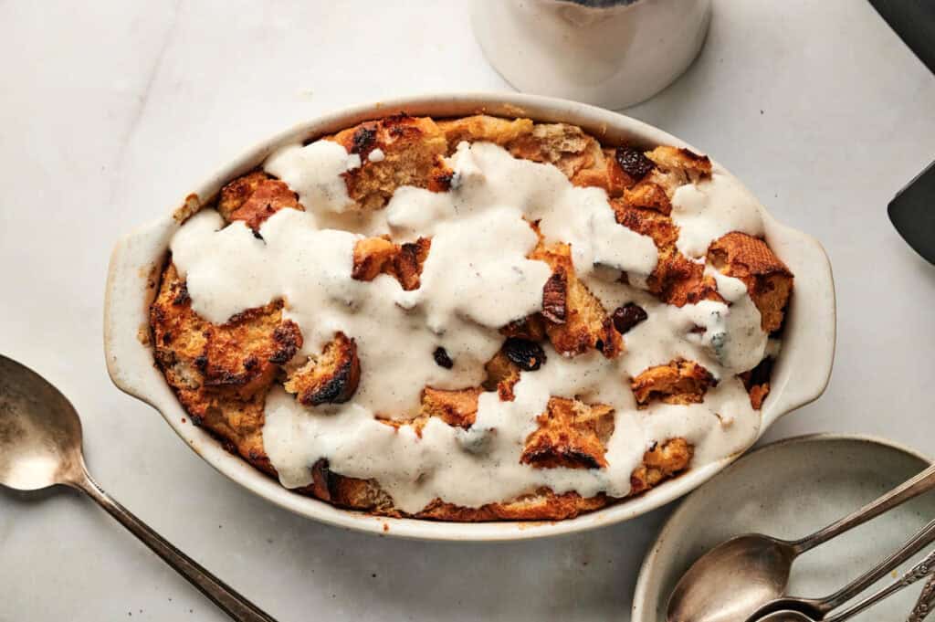 An oval dish brimming with air fryer bread pudding is perfectly topped with a luscious vanilla sauce. Two spoons and a small bowl accompany the dish, all elegantly arranged on a sleek marble surface.