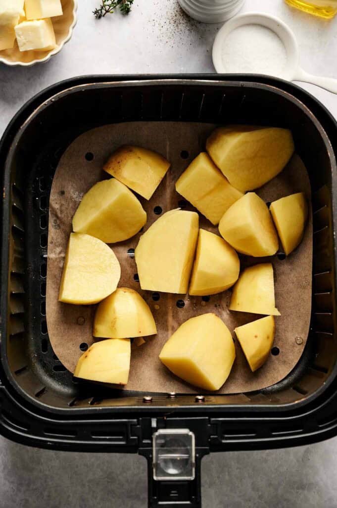 Chopped potatoes in an air fryer basket on parchment paper, with surrounding ingredients like salt, butter, herbs, and the promise of turning into creamy Air Fryer Mashed Potatoes.