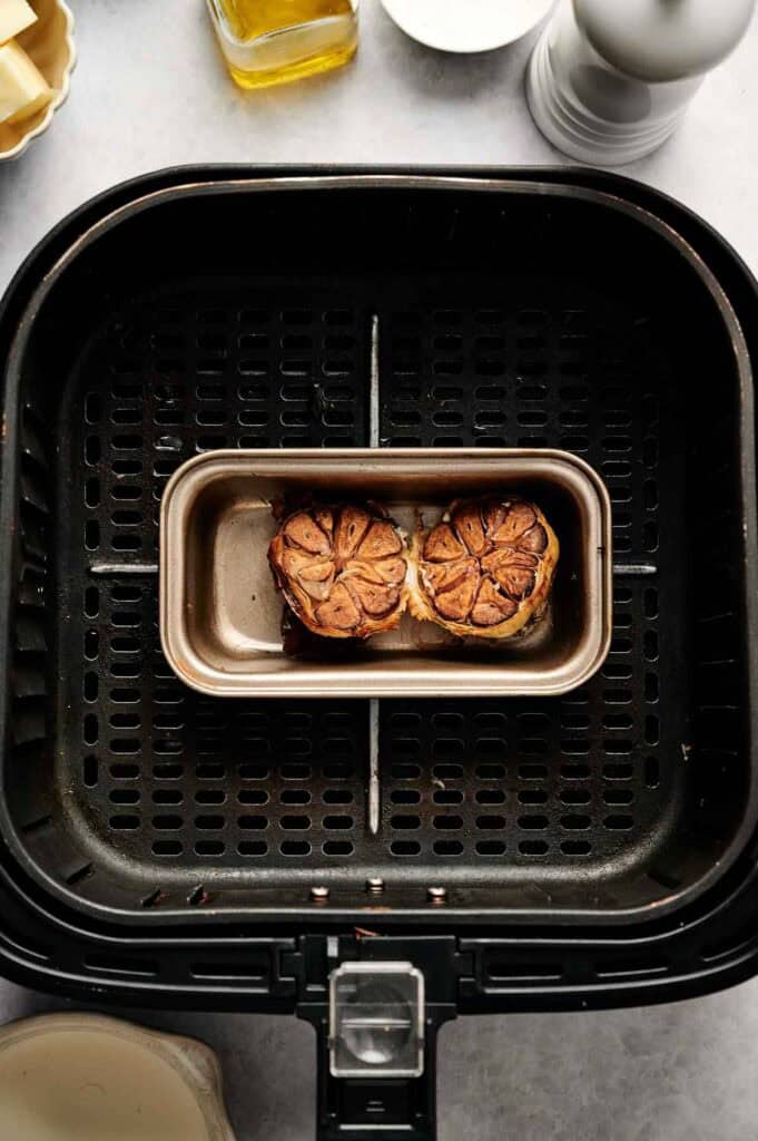 Roasted garlic bulbs nestle in a small tray inside an air fryer basket, with various kitchen items nearby, ready to create the perfect Air Fryer Mashed Potatoes.