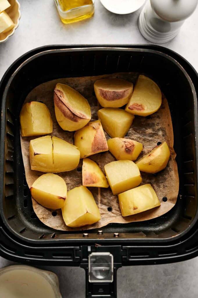 Potato wedges, soon to become crispy delights, are arranged on parchment paper in an air fryer basket, poised for cooking perfection. A pepper mill and olive oil bottle stand in the background, hinting at flavors to come. Ideal companions to air fryer mashed potatoes!.