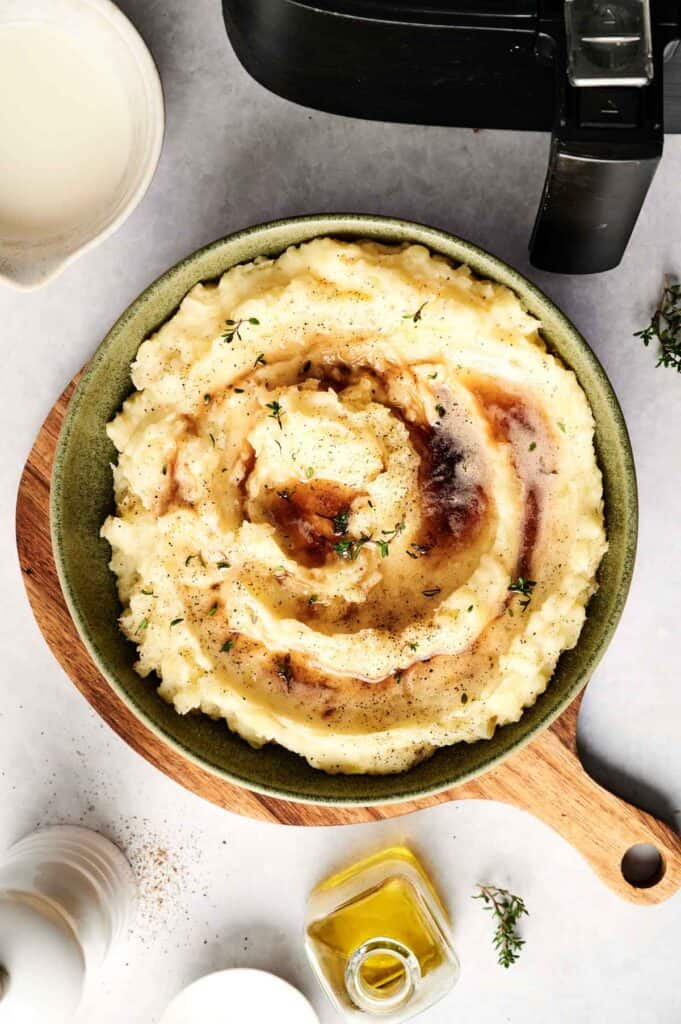A bowl of air fryer mashed potatoes with gravy on top, garnished with herbs, sits on a wooden board beside a small bottle of oil and a container.