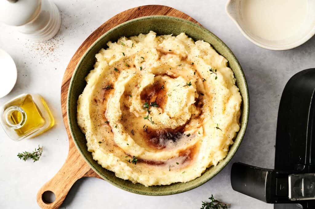 A bowl of air fryer mashed potatoes with brown gravy swirls, garnished with herbs, sits on a wooden board. Nearby are a jug of cream, a small bottle of oil, and a pepper grinder.