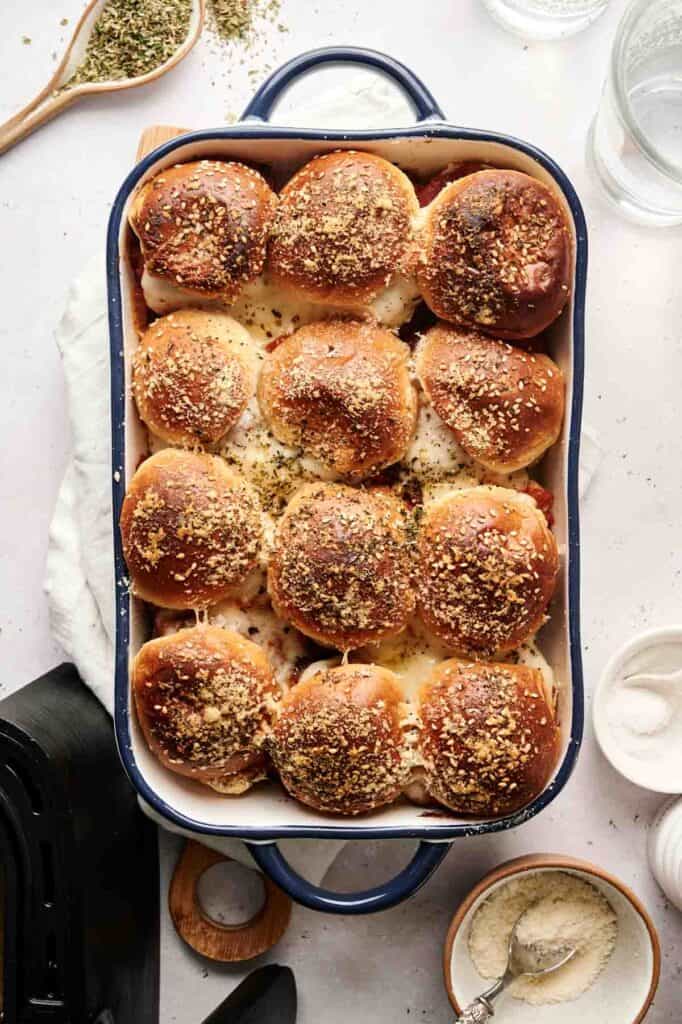 A baking dish filled with twelve browned, seasoned dinner rolls, reminiscent of air fryer meatball sliders, topped with herbs and grated cheese.