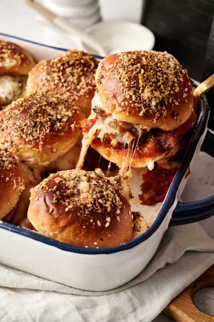 Air Fryer Meatball Sliders are baked in a dish with melted cheese and a crumb topping, one slider being lifted out.