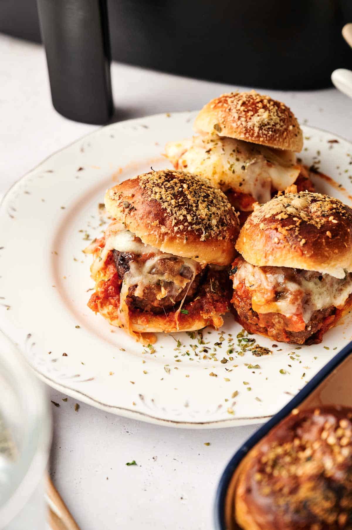 Three air fryer meatball sliders with tomato sauce and melted cheese rest on a white plate, garnished with fresh herbs.