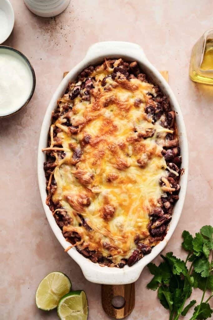 Oval baking dish filled with a baked, cheesy Beef Enchilada Casserole, surrounded by sliced lime, cilantro, a bowl of sour cream, and a bottle of oil on a light countertop.