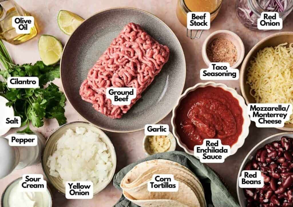 A flat lay of labeled taco ingredients for a Beef Enchilada Casserole, featuring ground beef, red and yellow onion, beef stock, taco seasoning, two cheeses, enchilada sauce, red beans, corn tortillas, sour cream, cilantro, olive oil, salt, pepper, and garlic.