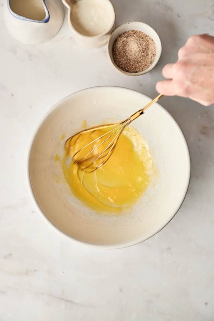 A hand whisking egg yolks in a white bowl on a marble surface, setting the stage for a delectable creation. Nearby are a ceramic pitcher, a bowl of cream, and spices, ready to enhance the flavors of an air fryer bread pudding masterpiece.