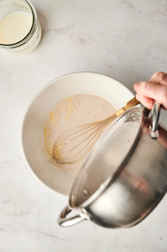 A person pours liquid from a pot into a mixing bowl with a whisk and light-colored batter, preparing the base for an air fryer bread pudding. Nearby, a glass jar filled with milk sits on a marble countertop, ready to add richness to the delightful creation.