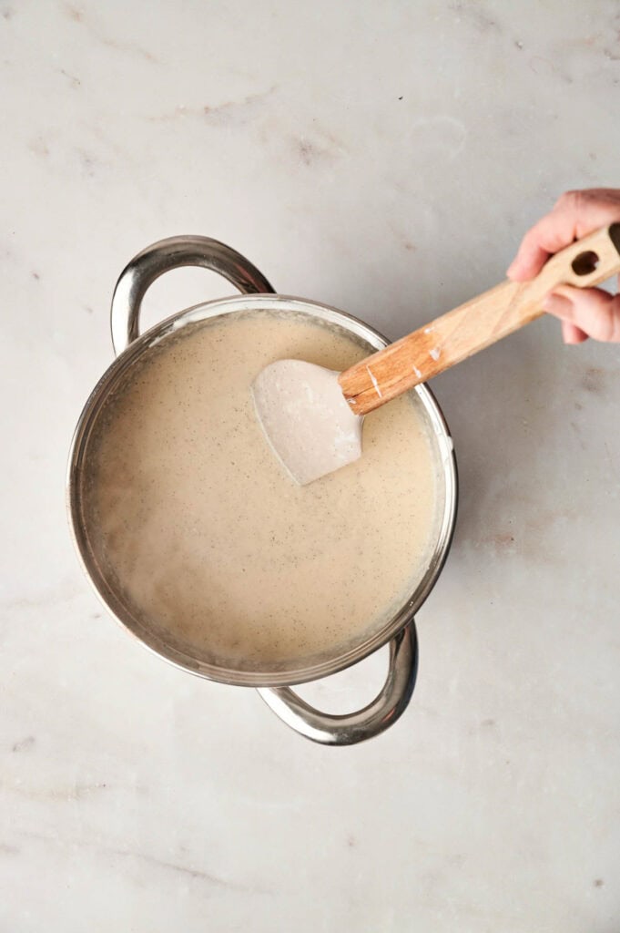Hand stirring a light beige mixture, reminiscent of creamy bread pudding, in a stainless steel pot with a wooden spatula atop a light marble surface&mdash;an inviting scene that echoes the warmth of freshly baked delights right out of an air fryer.