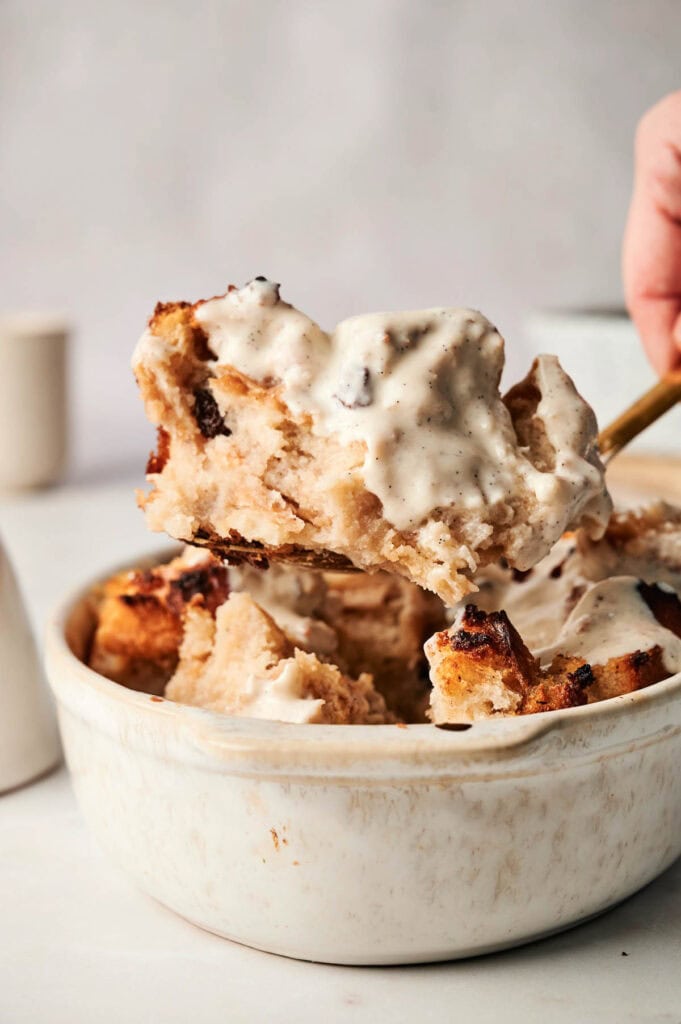 An air fryer bread pudding, nestled in a white ceramic bowl, is scooped up with a spoon, each bite dripping with creamy vanilla sauce.